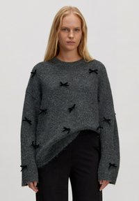 Grau gestrickter Pullover in lockerer Passform, der an der Vorderseite mit schwarzen Stoffschleifen sowie an Saum und Ärmeln mit rohen Kanten verziert ist.