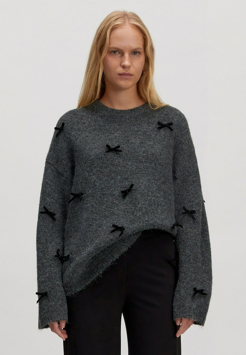 Grau gestrickter Pullover in lockerer Passform, der an der Vorderseite mit schwarzen Stoffschleifen sowie an Saum und Ärmeln mit rohen Kanten verziert ist.