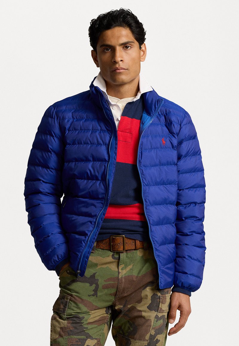 Homem usando uma jaqueta azul acolchoada sobre uma camisa listrada em vermelho e azul-marinho com colarinho branco e calças cargo em padrão de camuflagem.