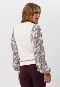 Witte sweatshirt met gemusterde bruine pofmouwen, zachte textuur, ronde halslijn en aansluitende taille, gecombineerd met bordeauxrode broek.