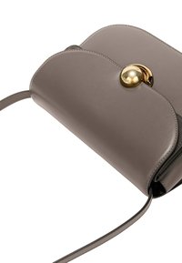 Borsa a tracolla compatta in pelle taupe con patta arrotondata, hardware circolare dorato e tracolla sottile e regolabile. Texture liscia con dettagli minimi.