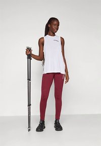 Valge ilma varrukateta topp musta logoga, marooni värvi retuusid õmblusdetailidega, mustad jalanõud, hoides musti matkakeppe, seistes heleda taustaga.