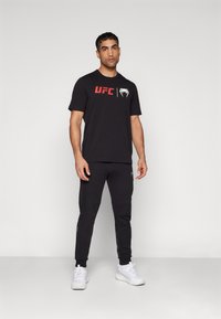 Sort bomulds t-shirt med rødt "UFC" logo og hvid grafik. Bæres med matchende sorte joggingbukser og hvide træningssko.