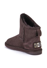 Botín de ante marrón con forro de pelo suave, punta redondeada y un acento metálico de marca en el lateral. Suela de goma plana.