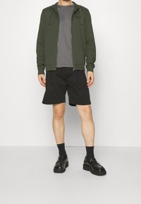 Felpa con cappuccio verde scuro con zip, T-shirt grigia, pantaloncini neri e mocassini neri. La felpa ha coulisse e dettaglio con logo. Outfit semplice e casual.