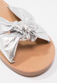 Sandale en métal argenté avec un dessus en tissu noué, semelle intérieure texturée marron et détail de semelle beige piqué.