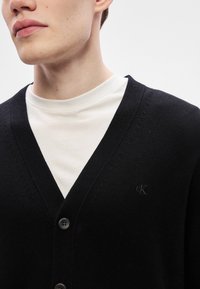 Cardigã preto de malha com decote em V, com dois botões e um logótipo em relevo. Usado sobre uma t-shirt branca de gola redonda.