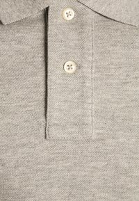 Šedé polo tričko s texturovanou pleteninou, tří knoflíkovou légou a klasickým límcem. Knoflíky jsou světlé a mají čtyři dírky.