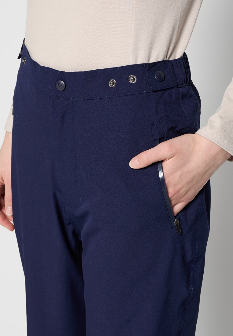 Person, der eine marineblaue Hose mit Druckknöpfen und einer mit Reißverschluss versehenen Seitentasche trägt, mit der Hand in der Tasche.