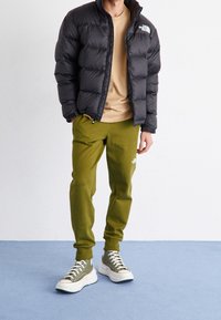 Person som bär en svart pufferjacka, beige skjorta, olivgröna träningsbyxor och matchande hög-toppsneakers som står på ett blått golv.