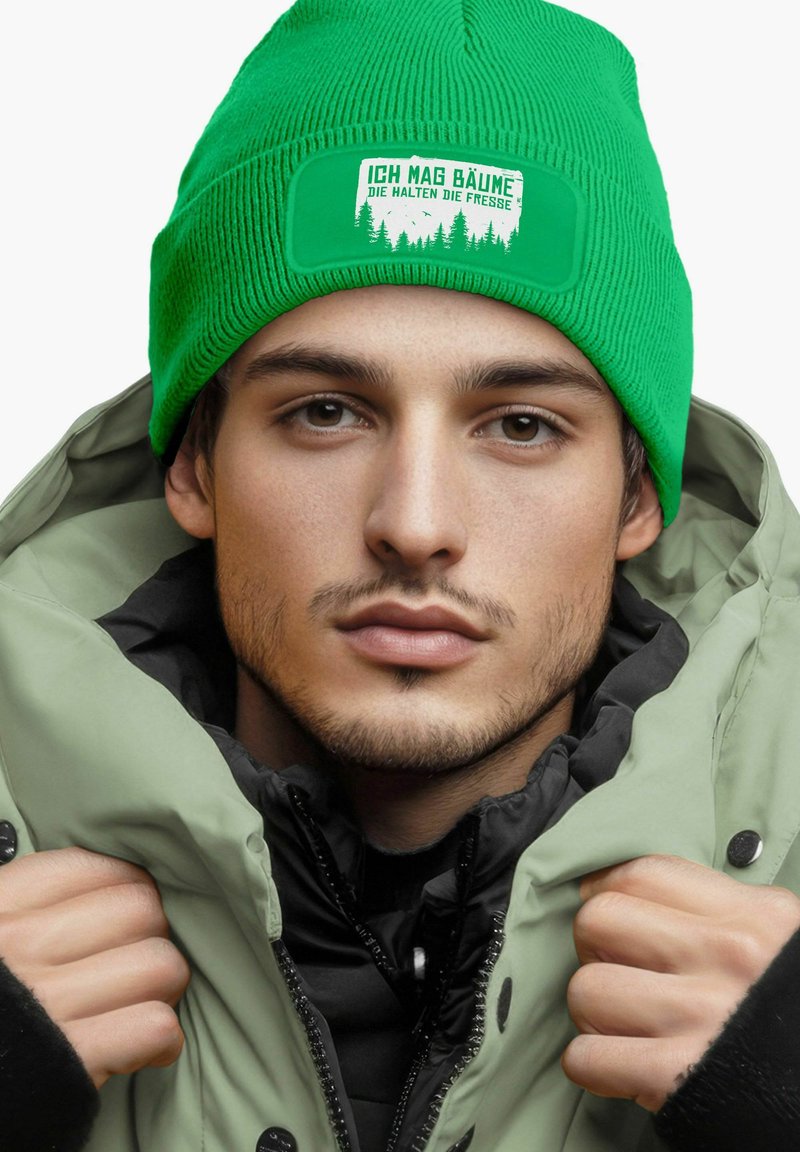 Green knitted beanie with a white patch featuring the text "Ich mag Bäume." Worn with a layered gray and black winter jacket.