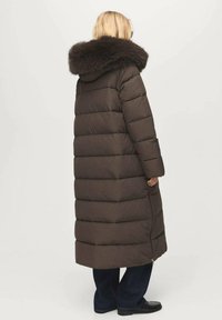 Abrigo acolchado largo marrón con capucha forrada de piel, que presenta un diseño acolchado y cremallera lateral, llevado sobre pantalones azul oscuro y zapatos.