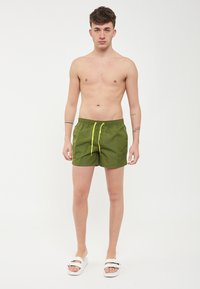 Shorts de bain verts avec un cordon de serrage à la taille, ornés de accents jaunes fluo ; matière légère ; longueur au-dessus du genou ; design simple.