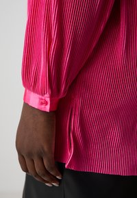 Blusa fucsia con plisado, de manga larga, con puños de satén y aberturas laterales. Presenta una superficie texturizada con un acabado brillante.
