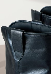 Geantă din piele neagră cu textură netedă, colțuri superioare rotunjite și mânere laterale rezistente. Cusături vizibile și detalii de design minimalist.