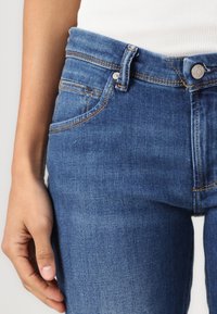 Keskmise sinise värviga denim teksad, millel on sile viimistlus, viie tasku disain, vaskvärvi õmblused ja metallnupp vöökoht. 
