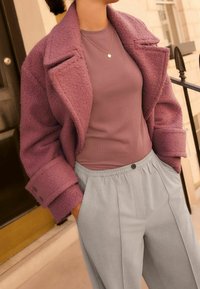 Manteau oversize texturé violet, porté sur un haut moulant mauve, associé à un pantalon gris clair à taille élastique avec plis sur le devant et poches.