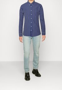 Uomo che indossa una camicia a righe blu navy e bianca con bottoni, jeans blu chiaro e stivali in pelle nera con lacci, in piedi contro uno sfondo bianco uniforme.