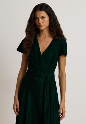 Femme aux longs cheveux bouclés portant une robe portefeuille à manches courtes vert foncé, nouée à la taille, debout devant un fond clair uni.