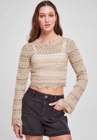 Beige hekletrøye med åpenmønster, bølgete kanter og vide ermer, kombinert med mørke denim høytlivsshorts.