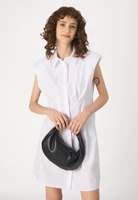 Λευκό φόρεμα πουκάμισου με κοντά μανίκια, κουμπωτό μπροστινό μέρος και καμπυλωτό στρίφωμα; το μοντέλο κρατά μια μαύρη δερμάτινη τσάντα hobo. Λείος υφασμάτινος σχεδιασμός, μοντέρνα εμφάνιση.