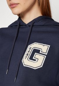 Felpa con cappuccio blu navy con un emblema in pile a forma esagonale che presenta la lettera "G" e la parola "GANT" ricamate in colori contrastanti.