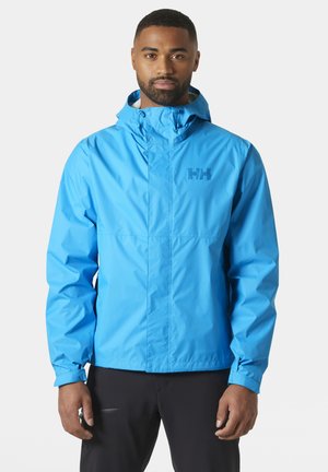 Man draagt een helderblauwe, waterdichte jas met capuchon, rits en drukknoopsluiting, en zwarte broek, staand tegen een effen achtergrond.