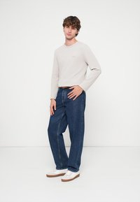 Ανοιχτό γκρι μακρυμάνικο πουκάμισο με στρογγυλή λαιμόκοψη, συνδυασμένο με αέρινα μπλε τζιν και λευκά αθλητικά παπούτσια με καφέ σόλες. Απλός σχεδιασμός, άνετη εφαρμογή.