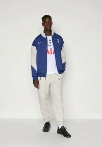 Mann in blauer und weißer Tottenham Hotspur Jacke, weißem Shirt mit Logo, hellbeigen Jogginghosen und schwarzen Sneakers, der vor einem weißen Hintergrund steht.