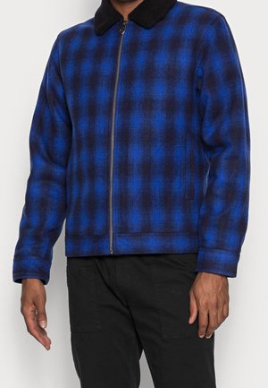 Homme portant une veste zippée à carreaux bleus et noirs avec un col noir, associée à un pantalon noir.