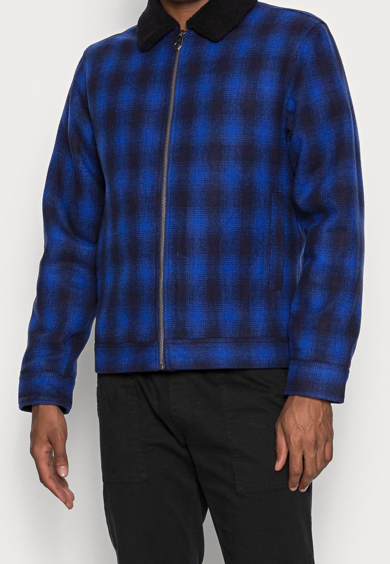 Homme portant une veste zippée à carreaux bleus et noirs avec un col noir, associée à un pantalon noir.