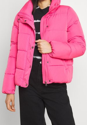 Femme portant une veste matelassée rose vif par-dessus un haut rayé noir et blanc et un pantalon noir taille haute, main ajustant la fermeture éclair de la veste.