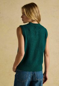 Zaļš bezdelīgas piedurknes adīts džemperis ar apaļu kaklu un ribotu apdari, kombinēts ar ziliem džinsiem. Tekstūra izskatās mīksta un ērta.