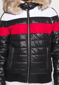 Svart pufferjacka med ett vitt och rött horisontellt rand. Har en fuskpäls-fodrad huva, dragkedjestängning och sidofickor.