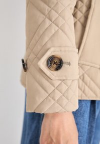 Beige quilted jacket sleeve featuring a hexagonal cuff with a button. Material is smooth with contrasting stitching and a decorative button detail.