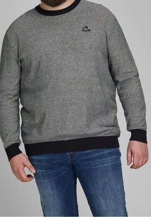 Mężczyzna ubrany w szary i czarny sweter dzianinowy z małym logo na piersi oraz niebieskie dżinsy, stojący na tle jednolitego szarego tła.