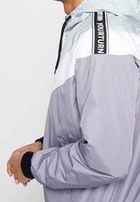 Lättvikts, färgblockerad vindjacka i grått och vitt. Har en huva, svarta accenter på ärmarna och en dragkedja framtill för enkel påtagning.