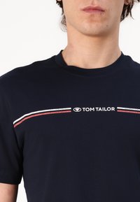 Mørkeblå T-skjorte laget av bomull, med rund hals, korte ermer, og et horisontalt design med hvite og røde striper og logo.