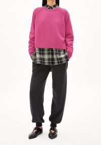 Pull fuchsia superposé sur une chemise à carreaux noir et blanc, associé à un pantalon gris foncé fuselé et des chaussures noires à bride.