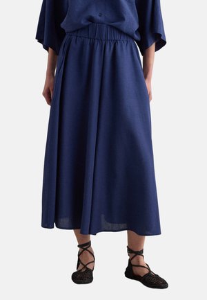 Mujer con falda midi azul marino con cintura elástica y zapatos planos negros tejidos con correas en el tobillo, de pie frente a un fondo liso.