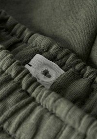 Tessuto verde con texture a pieghe, caratterizzato da una linguetta in tessuto bianco e un bottone trasparente. Il dettaglio evidenzia la cucitura e la morbidezza del materiale.