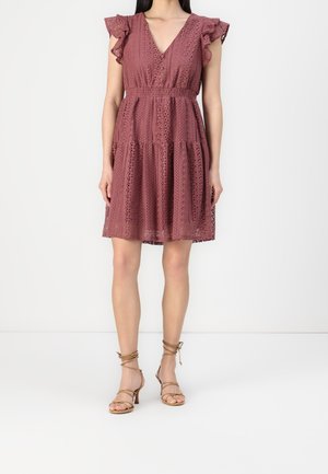 Kobieta ubrana w koronkową sukienkę do kolan w kolorze przybrudzonego różu, z falbankowymi krótkimi rękawkami, nosząca beżowe sandały na wysokim obcasie ze paskami, stojąca na białej podłodze.