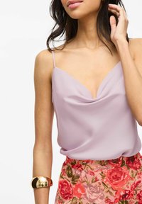 Camisola di raso lavanda con scollatura drappeggiata, abbinata a una gonna rosa floreale. Un bracciale dorato spesso completa l'outfit.