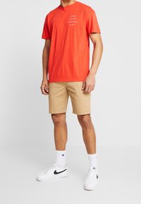 Man in oranje grafisch t-shirt, beige shorts, witte Nike sneakers en witte crew-sokken staand tegen een effen achtergrond.