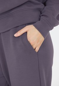 Mörklila sweatshirttyg med en mjuk textur, utrustad med en sidoficka och muddade ärmar. Designen är avslappnad och bekväm.