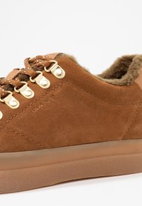 Baskets en daim marron avec des œillets dorés et des lacets beiges, dotées d'une doublure intérieure douce et moelleuse et d'une semelle en caoutchouc texturée.