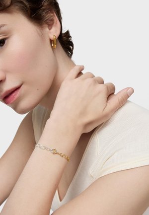 Pendiente de aro de oro, pulsera de plata y oro con detalle de cierre, llevados con una prenda de color claro. Texturas suaves con acabados metálicos contrastantes.