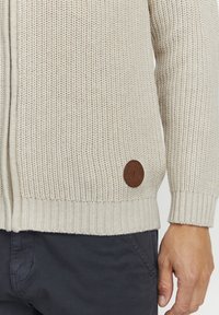 Cardigan de punto en beige claro con un patrón texturizado, puños y dobladillo acanalados, y un parche de cuero marrón en el lado inferior.