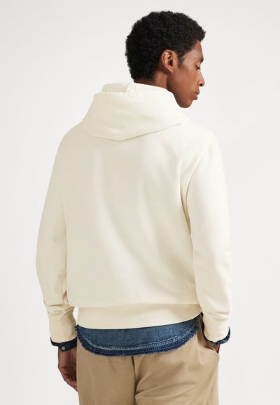 Vyras, stovintis nugara, dėvi kreminę džemperio su gobtuvu virš mėlynos spalvos džinsinio marškinio su nutrintu kraštu ir smėlio spalvos kelnes, rankos kišenėse, priešais vientisą foną.