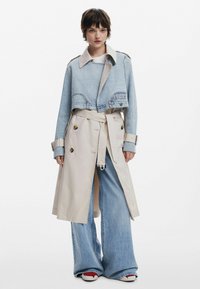 Abrigo de trinchera beige de mezclilla con un diseño corto. Presenta botones grandes, mangas anchas y pantalones de mezclilla acampanados. El calzado es rojo y blanco.